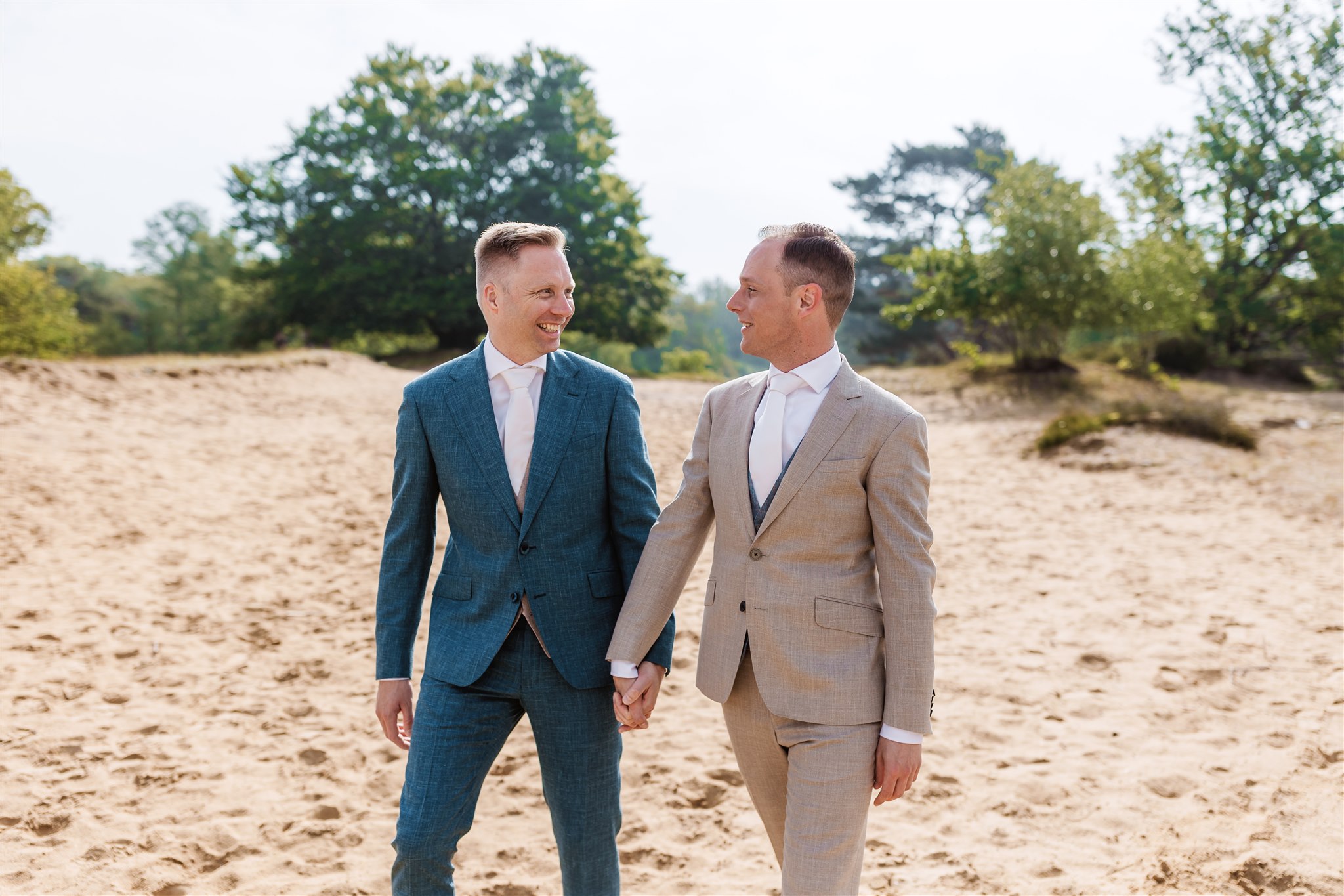 Twee mannen lopen tijdens het maken van trouwfoto's op hun bruiloft over een zandpad, lachend naar elkaar. De een draagt een blauw pak, de ander een beige pak.