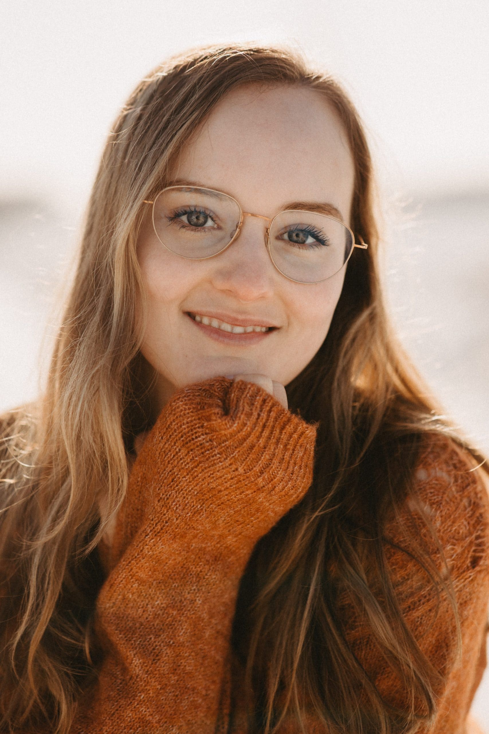 Een jonge vrouw met een bril en een oranje trui lacht naar de camera.