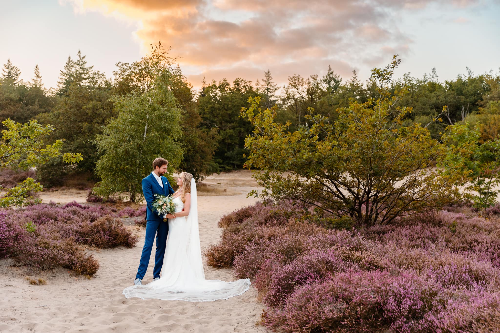 Bruidspaar kijkt elkaar aan op een trouwfoto tussen de bloeiende heide met prachtige ondergaande zon.