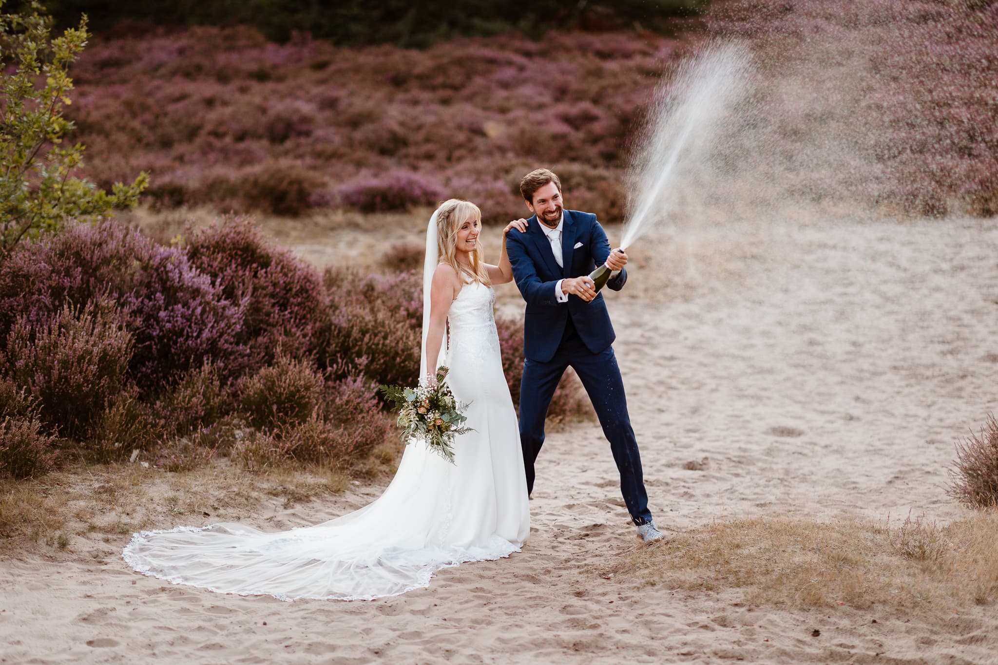 Bruidegom spuit met champagne terwijl zijn bruid heel hard lachend naast hem staat tijdens het maken van trouwfoto's.