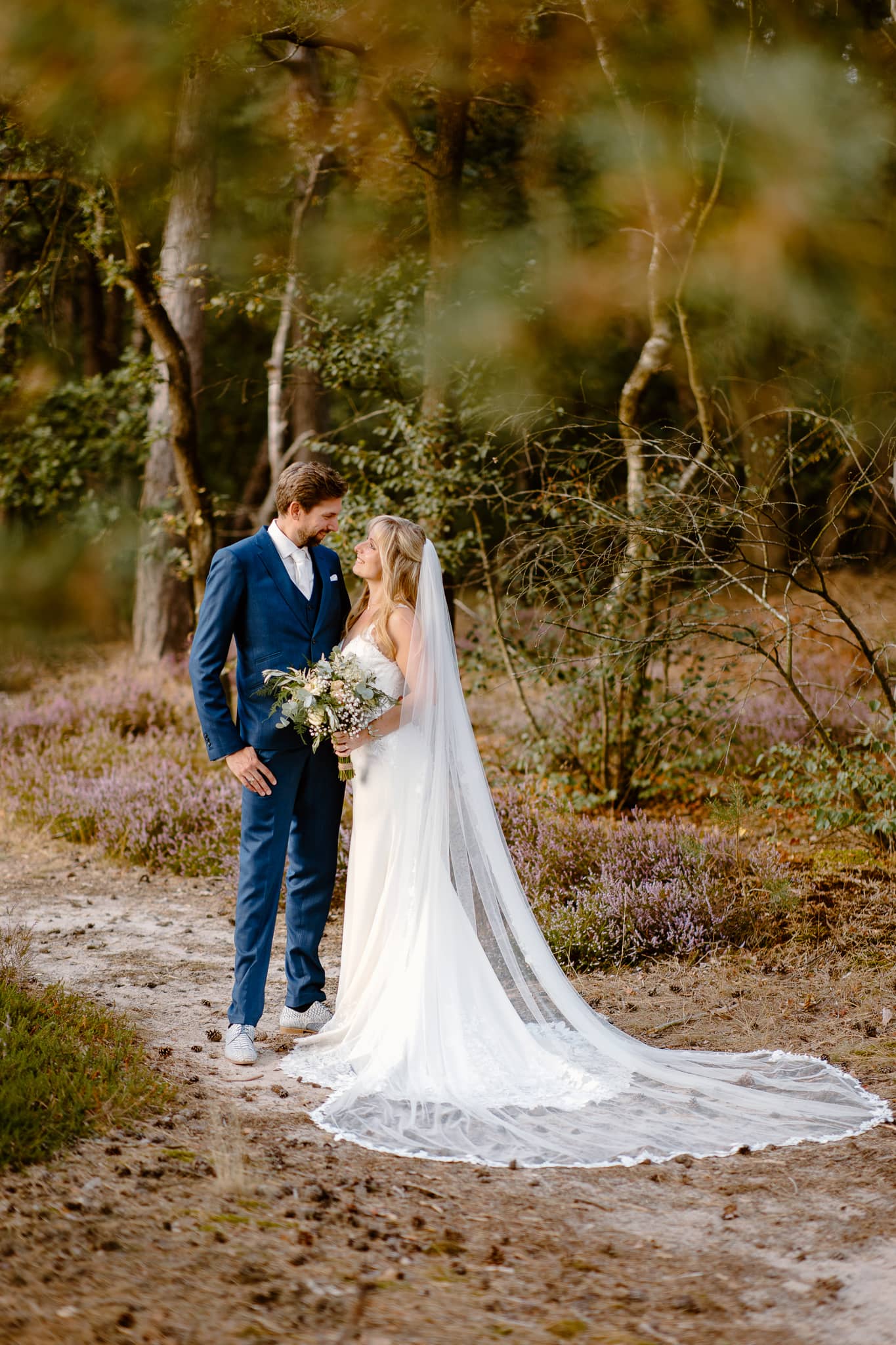 Bruid en bruidegom staan op de trouwfoto in het bos en kijken elkaar lachend aan.