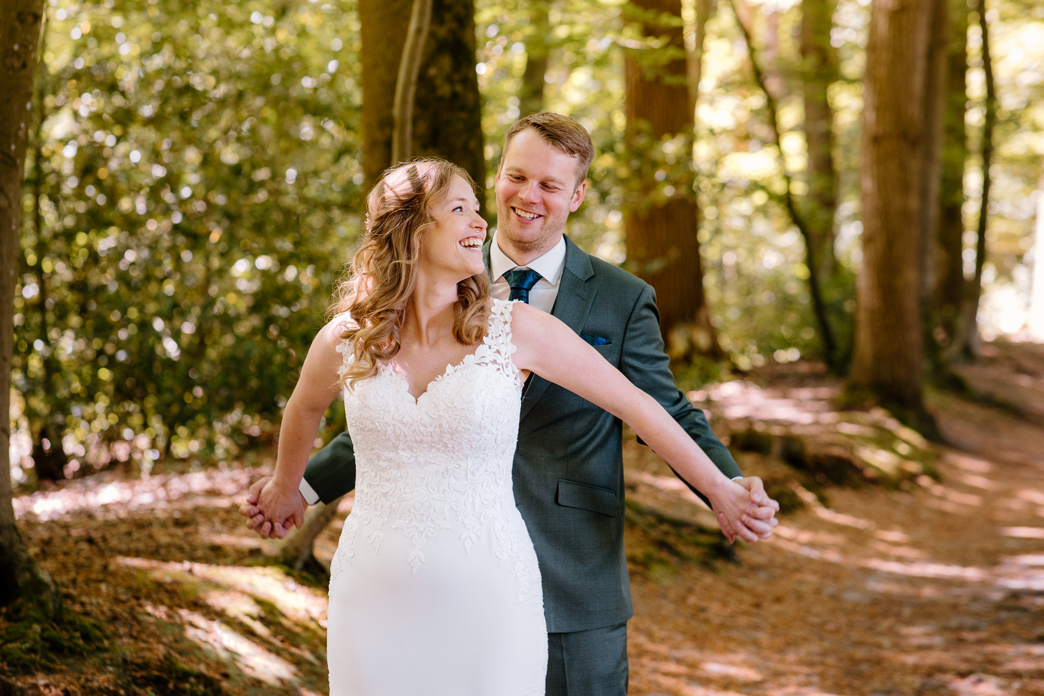 Een bruid en bruidegom kijken elkaar lachend aan tijdens het maken van hun trouwfoto's in het bos.