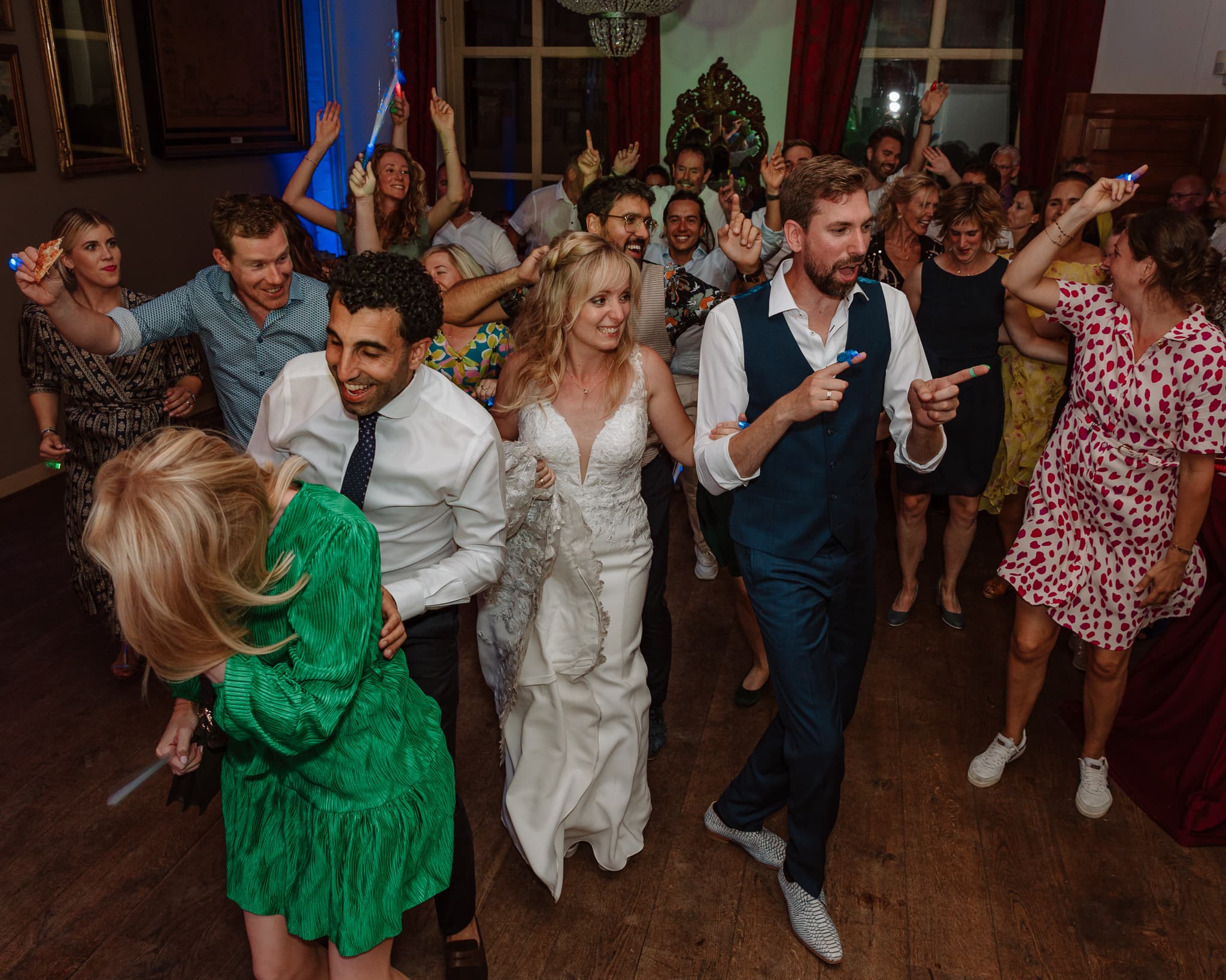 Bruidspaar en bruiloftsgasten dansen lachend in rijen van links naar rechts.