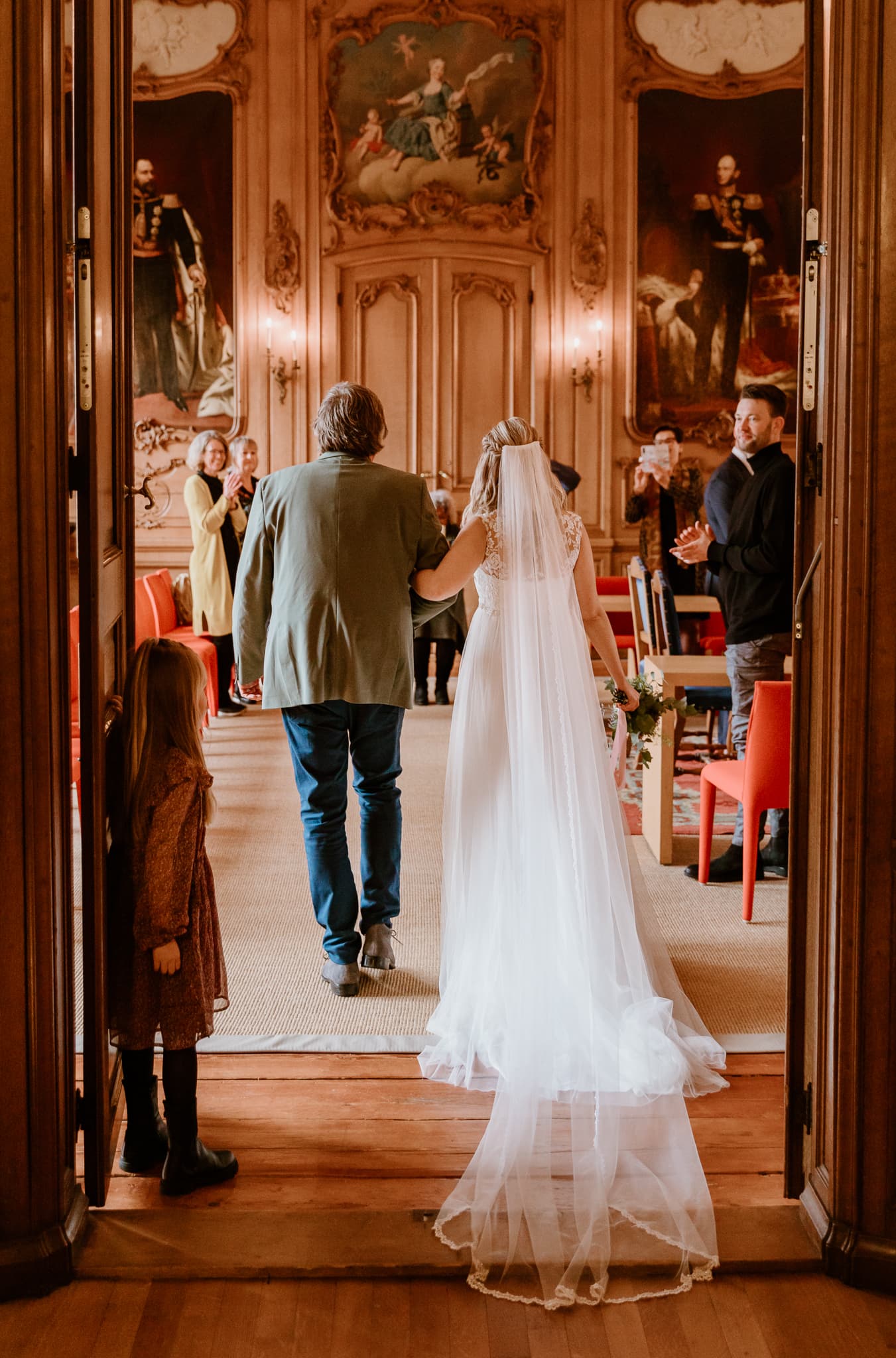 Bruid in witte jurk met lange sluier loopt gearmd met haar vader de trouwzaal van het stadhuis in Leeuwarden binnen.