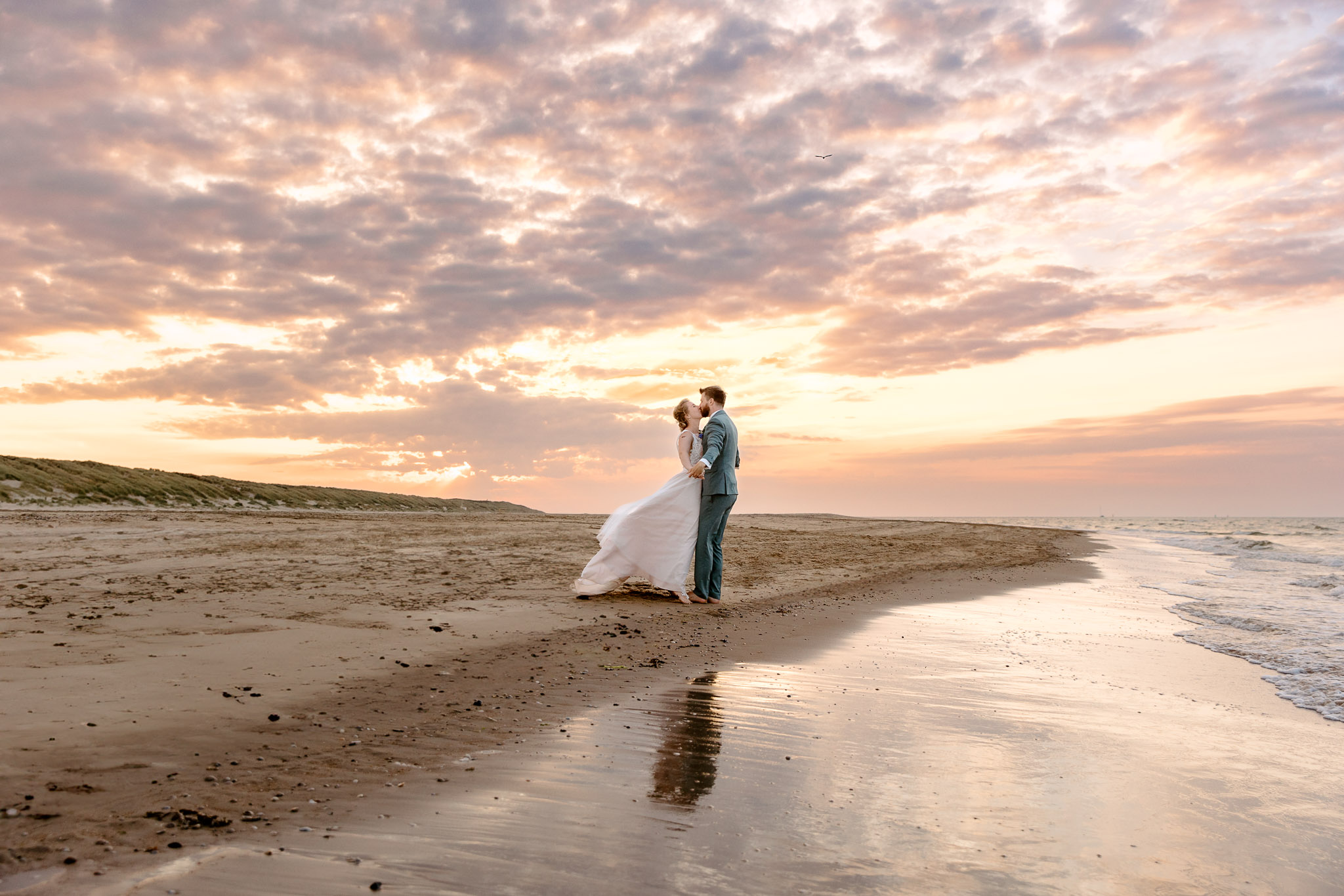 Een echtpaar viert hun strandbruiloft op Vlieland, terwijl ze bij zonsondergang op het strand staan en hun geloften uitspreken en ringen uitwisselen.