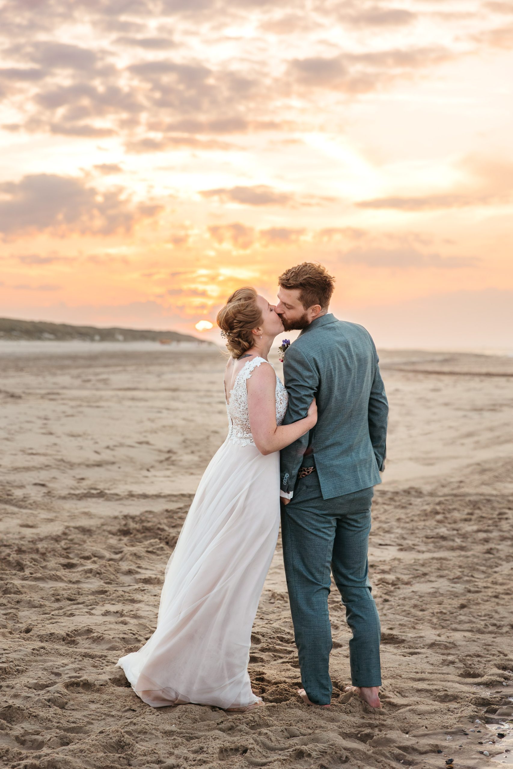 Een bruid en bruidegom kussen elkaar op het strand bij zonsondergang tijdens hun strandbruiloft op Vlieland.