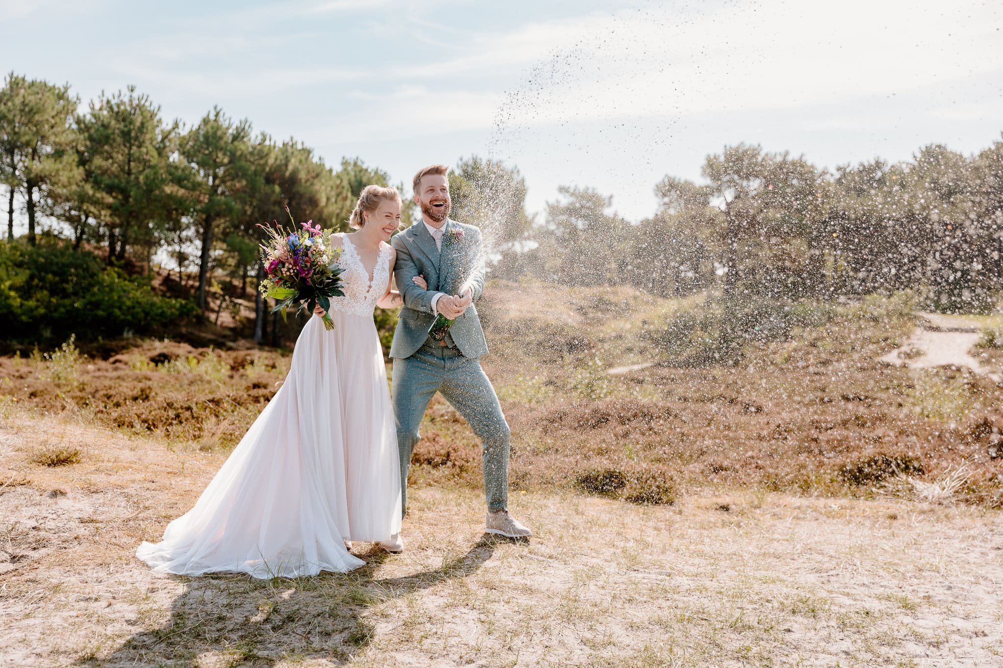 Een bruid en bruidegom die plezier hebben met het sproeien van water op elkaar tijdens hun Trouwen op Vlieland-feest in een veld.