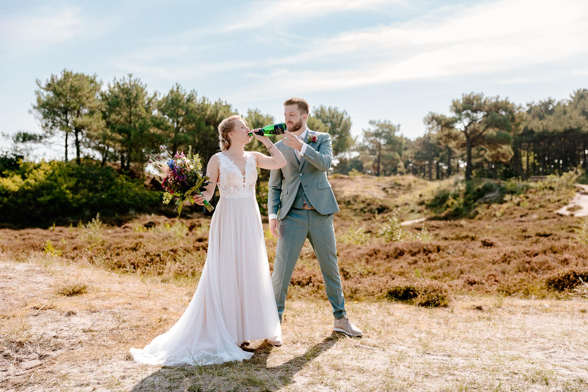 Een bruid en bruidegom genietend van een fles wijn tijdens hun Trouwen op Vlieland-ceremonie op een pittoresk veld.