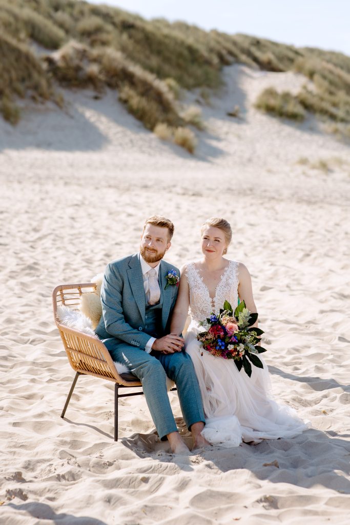 Een bruid en bruidegom die een romantische strandbruiloft hebben op Vlieland, zittend op een rieten stoel in het zand.