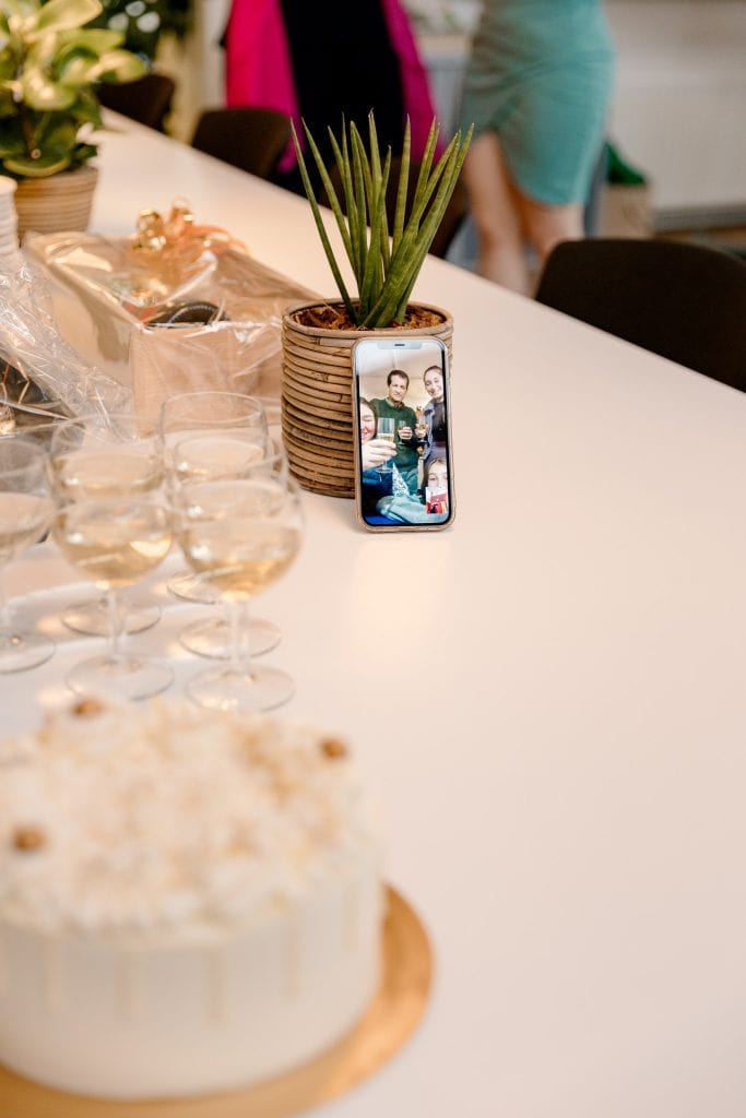 Een witte tafel met een taart erop en een telefoon waarop familie van de bruid vrolijk proost.