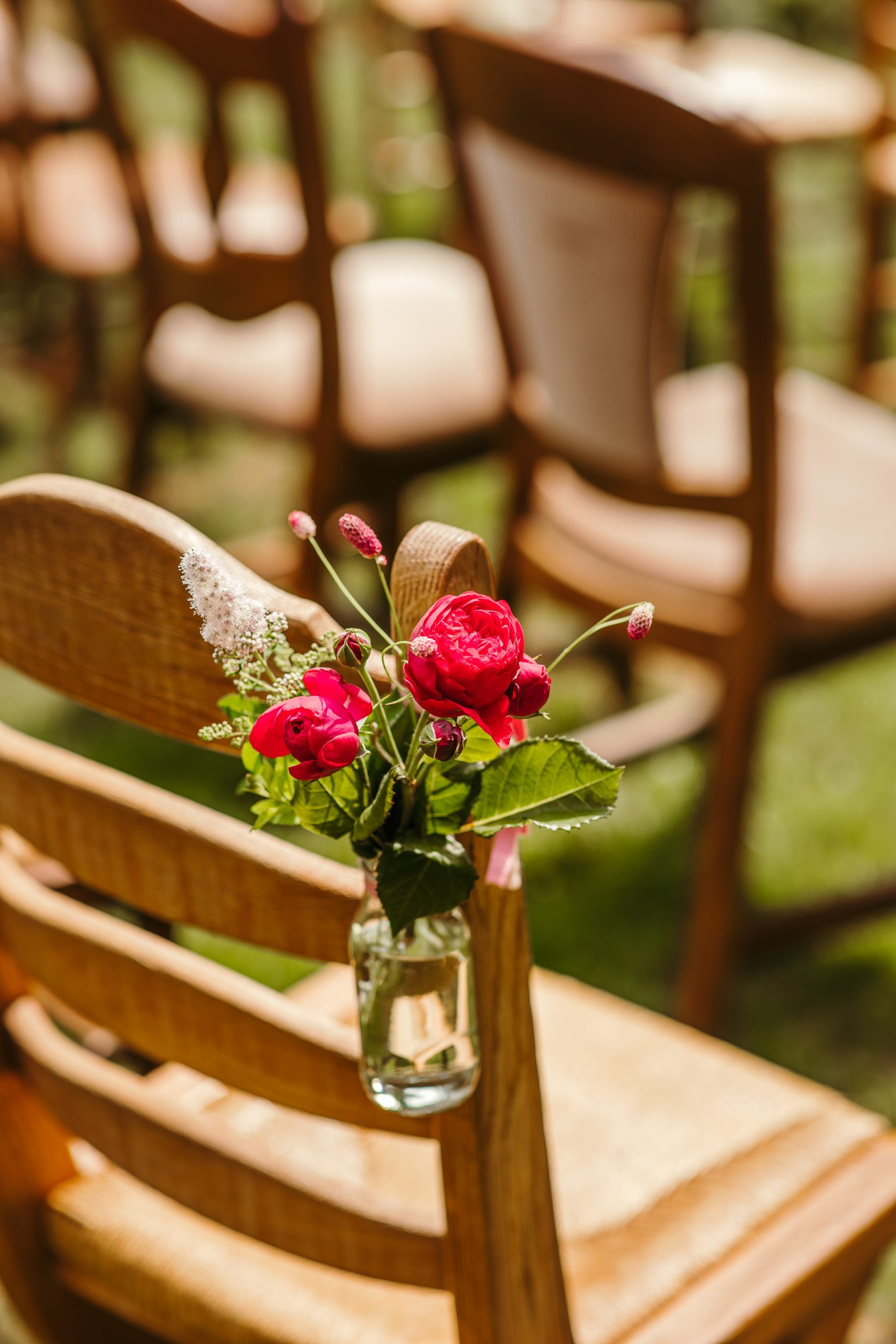 Een kleine glazen pot met levendige rode en witte bloemen is bevestigd aan de rugleuning van een rustieke houten stoel. De stoel maakt deel uit van een netjes opgestelde rij soortgelijke stoelen, waarschijnlijk opgesteld voor een buitenevenement of ceremonie op een zonnige dag.