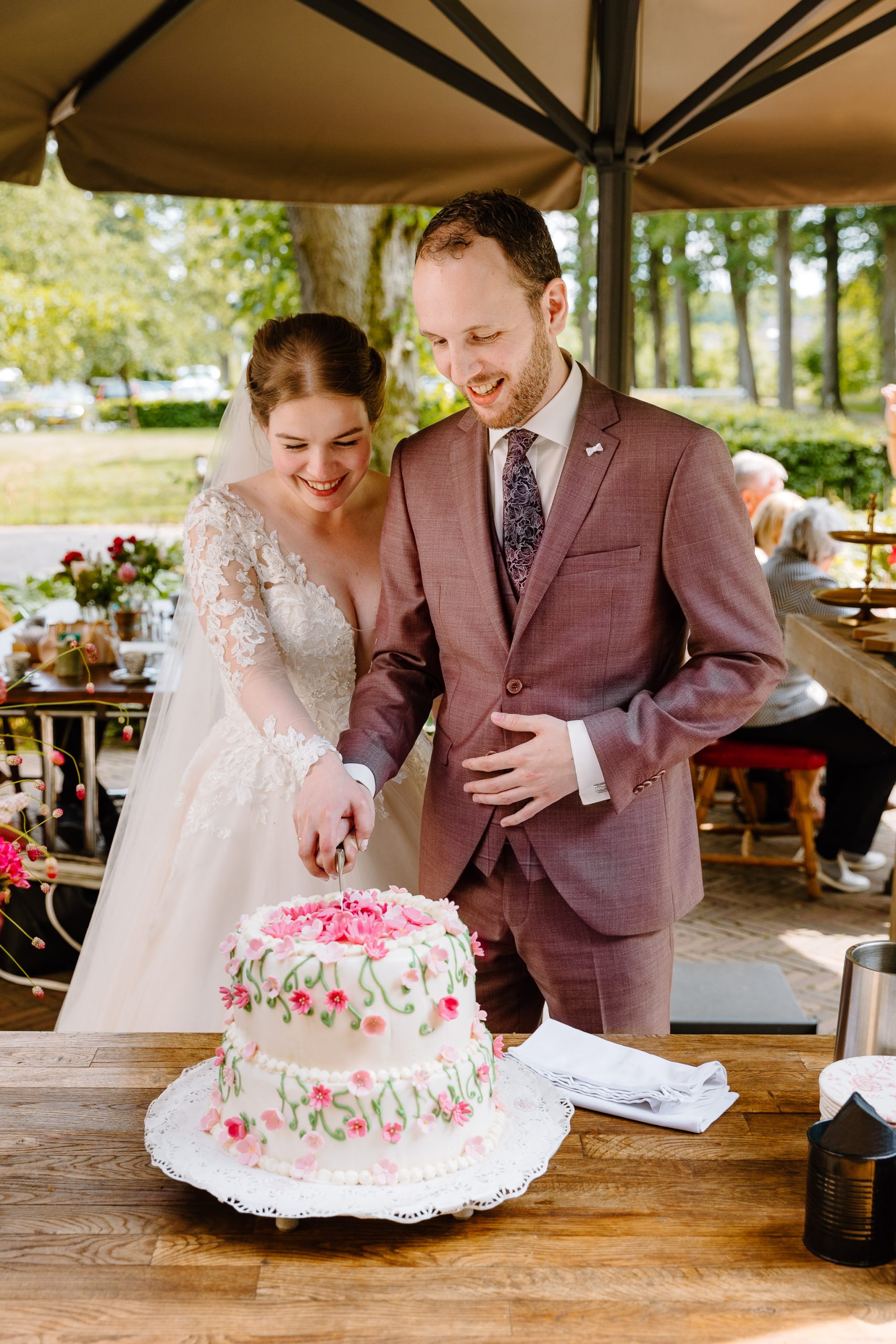 Een pasgetrouwd stel snijdt een witte bruidstaart versierd met roze bloemen. De bruid draagt een witte kanten bruidsjurk en de bruidegom draagt een kastanjebruin pak. Ze glimlachen en staan onder een baldakijn met groen op de achtergrond en gasten zitten vlakbij.