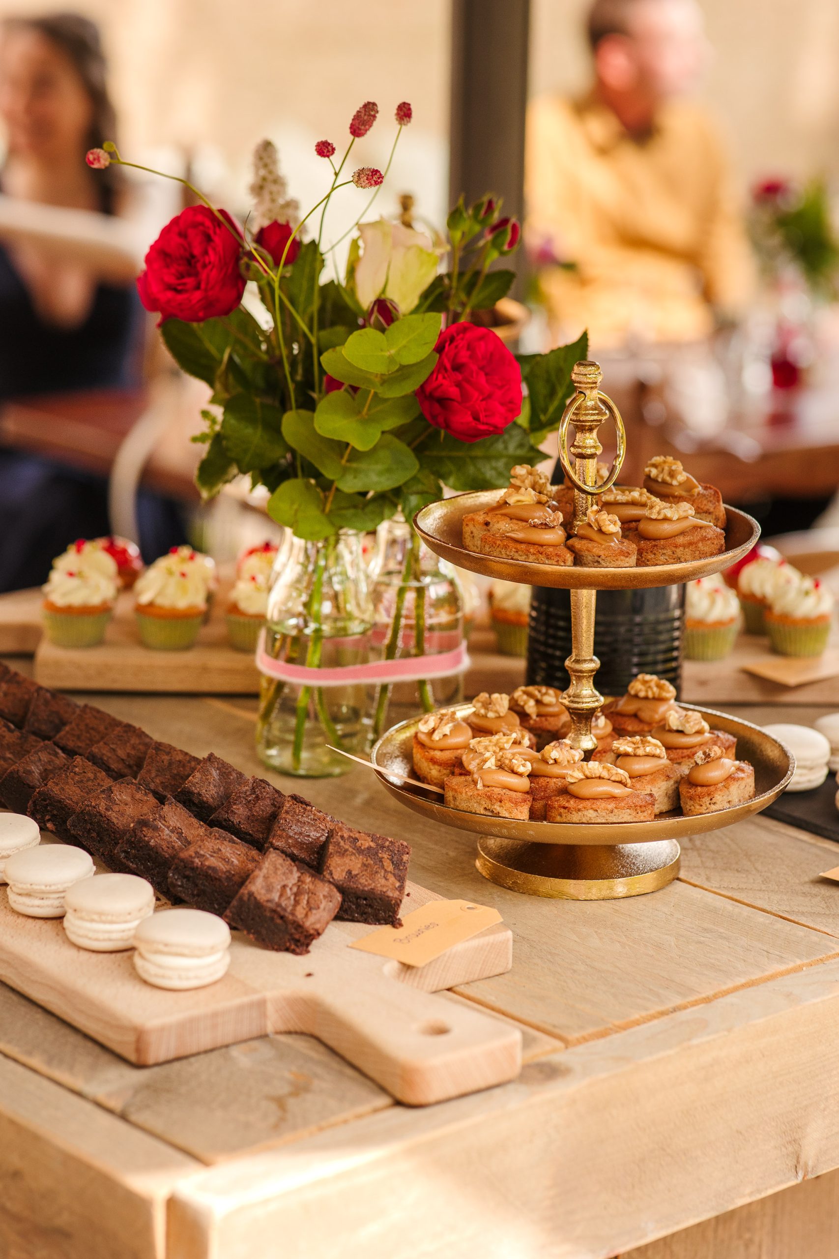 Een houten tafel versierd met een boeket rode rozen in een glazen vaas, omgeven door een assortiment bakproducten, waaronder brownies, macarons, cupcakes met witte en aardbeienglazuur en gebak met walnoten op een standaard met twee niveaus. Onscherpe achtergrond van mensen die dineren.