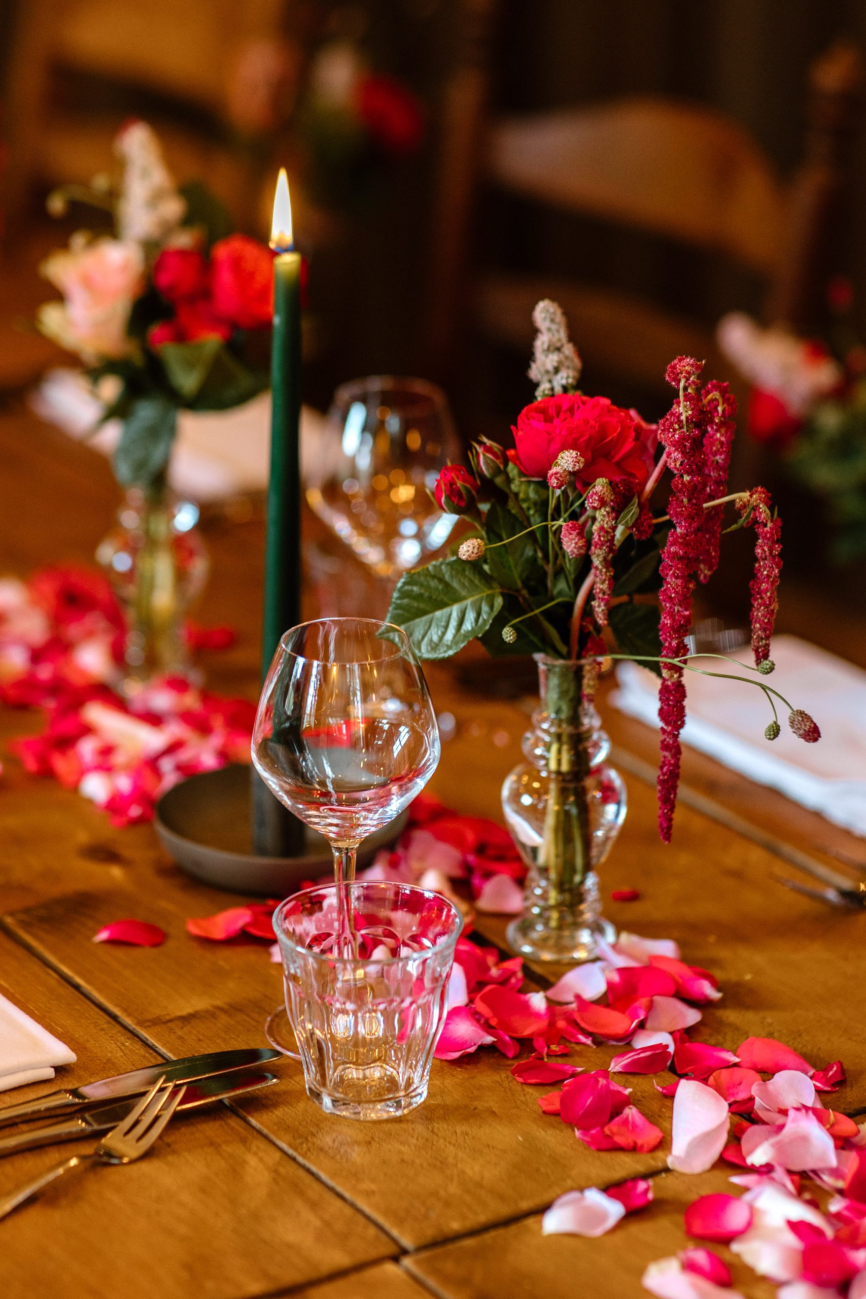 Een houten eettafel is elegant gedekt met lege wijnglazen, een brandende donkergroene kaars en vazen gevuld met levendige rode en roze bloemen. Rozenblaadjes liggen verspreid over de tafel en geven een romantisch tintje aan de setting. Bestek en servetten worden netjes geplaatst.