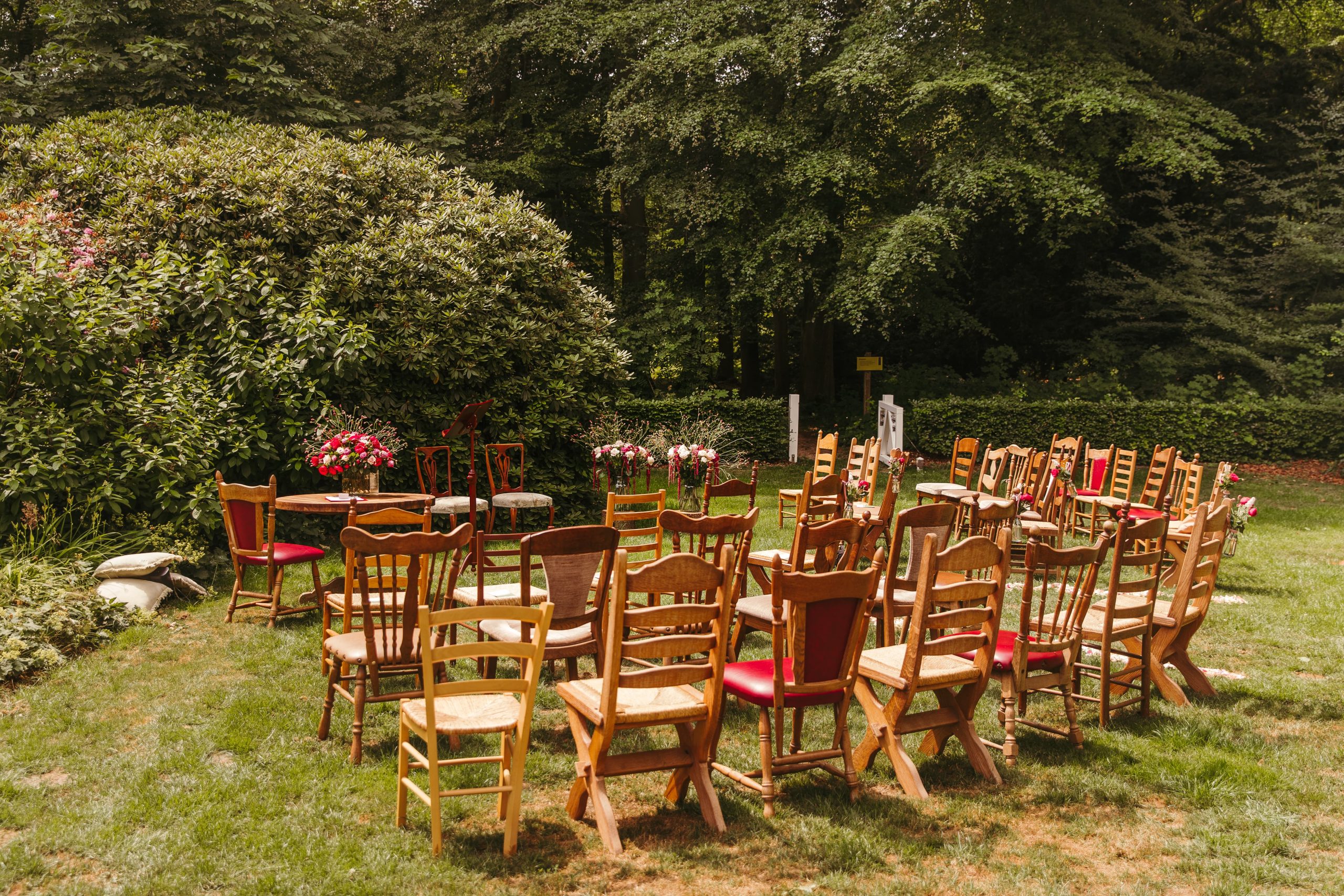 Een serene buitenomgeving met een cirkelvormige opstelling van vintage houten stoelen op een grasveld. Het gebied is omgeven door weelderige bomen en groen, met een paar kleurrijke bloemstukken op kleine tafels tussen de stoelen, wat doet denken aan een bruiloft of bijeenkomst.