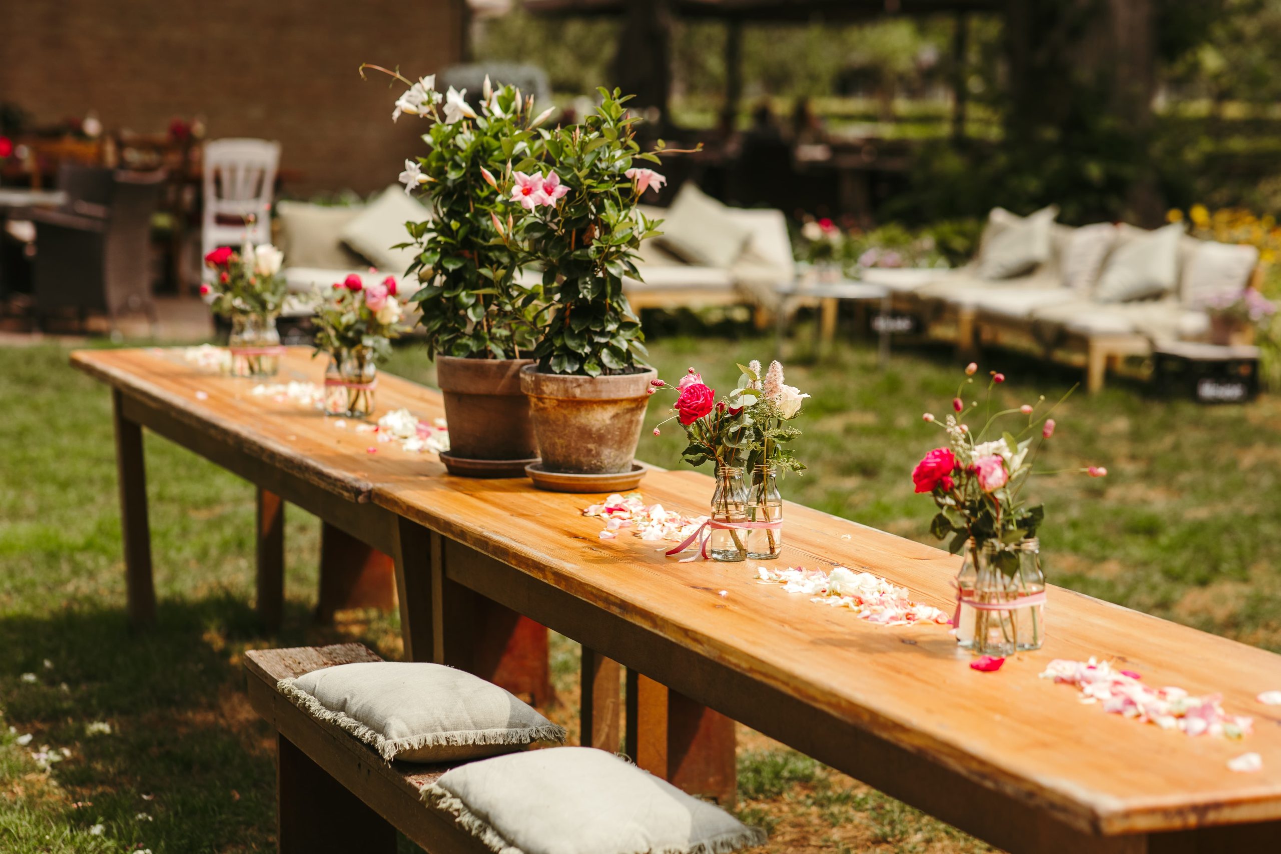 Een lange houten tafel buiten op een grasveld is versierd met bloemstukken in vazen en bloemblaadjes. Langs de tafel staan banken met kussens en op de achtergrond is onder een baldakijn een tuin met zitjes en groen zichtbaar.