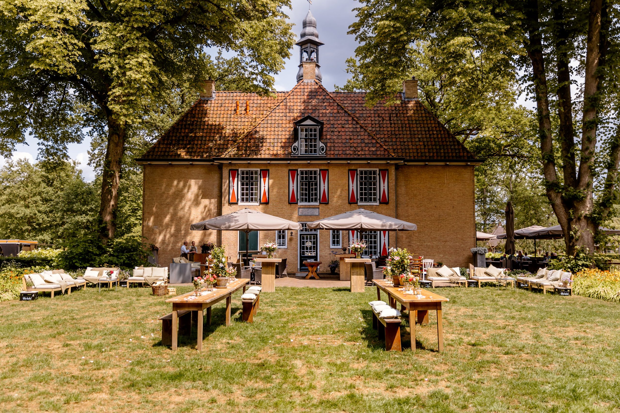 Er wordt een buitenevenement georganiseerd op een grasveld voor een groot bakstenen gebouw met een steil dak met dakspanen. Op het gras staan lange houten tafels en banken, versierd met bloemrijke middenstukken. De opstelling is voorzien van parasols voor schaduw en zitjes aan weerszijden.