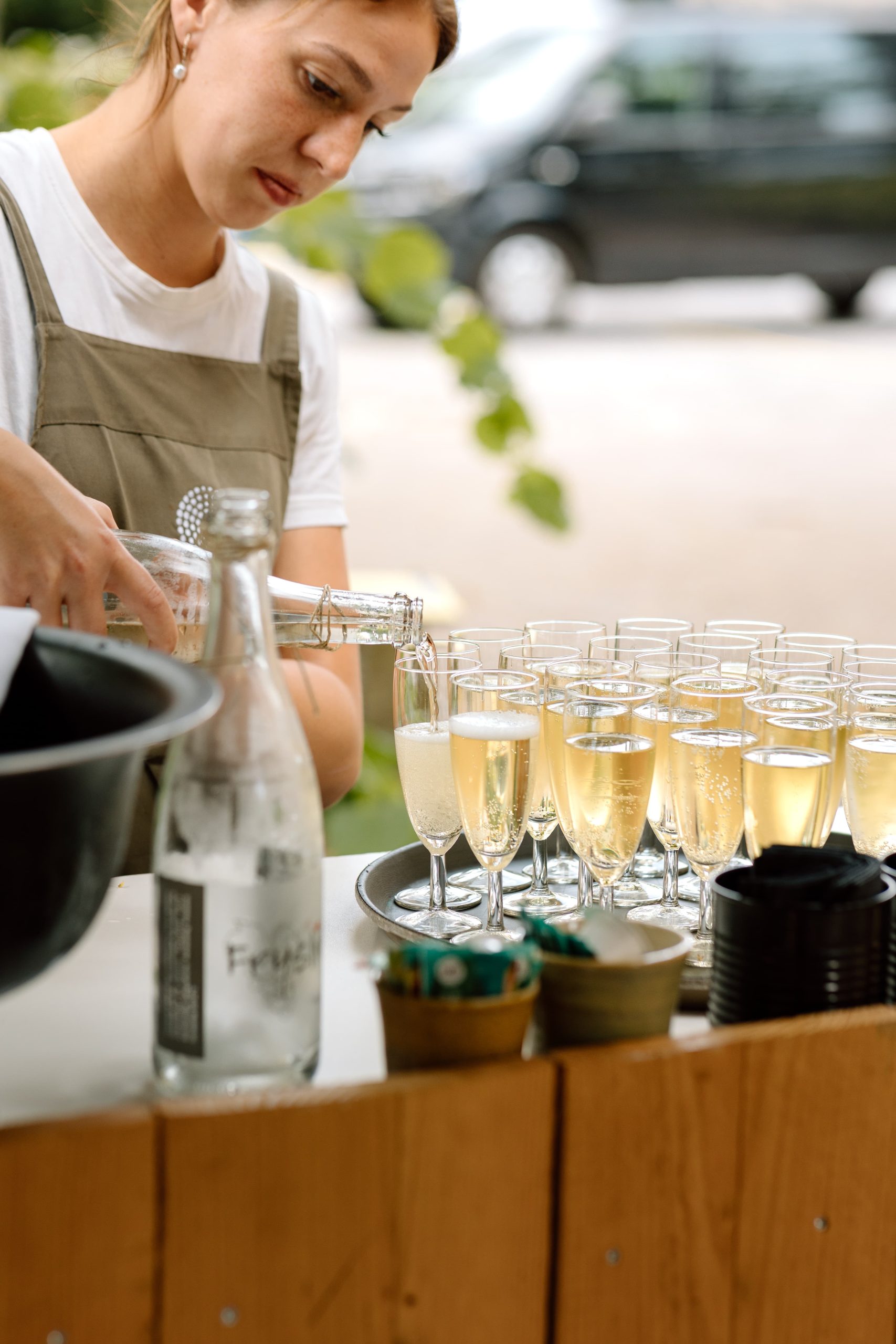 Een persoon die een schort draagt, giet champagne in fluitglazen die op een dienblad zijn geplaatst. De opstelling bestaat uit flessen, een emmer en kleine containers op een houten toonbank, met een onscherpe achtergrond van een straat en groen.