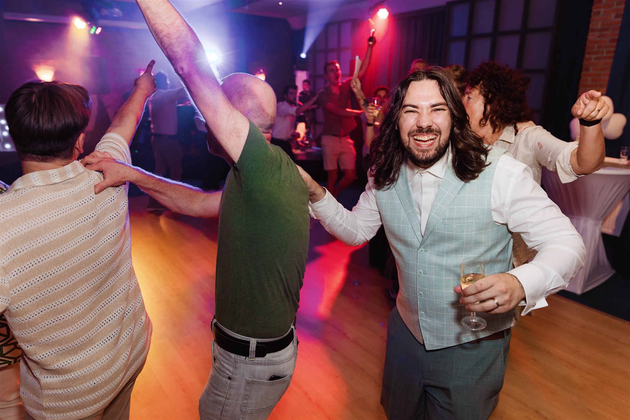 Een bruidegom kijkt breed lachend de camera in terwijl hij tijdens zijn trouwfeest de polonaise loopt.