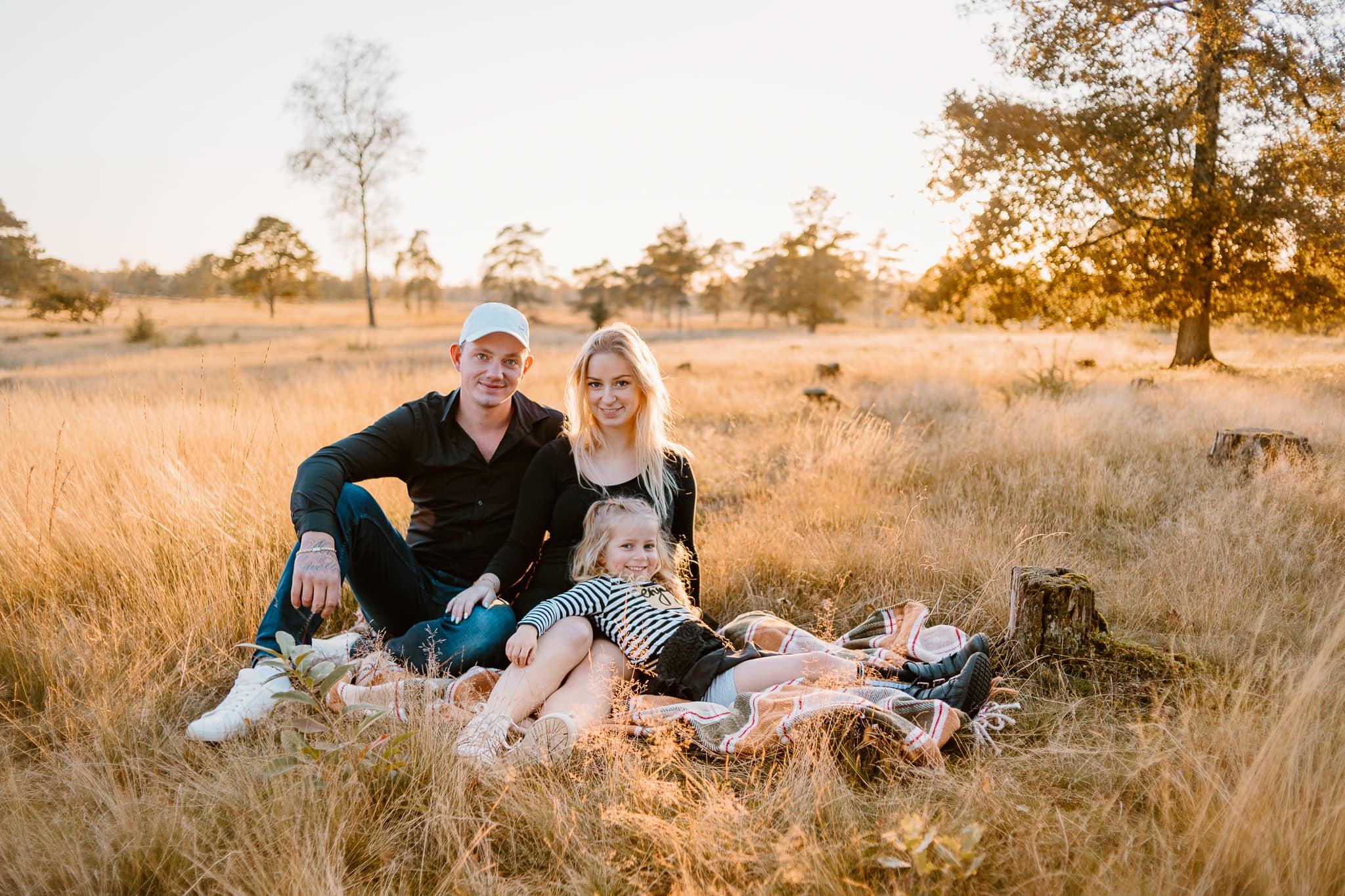 Jonge ouders met hun dochter van vier zitten tijdens zonsondergang op een kleed tussen de hoge gele grassen.