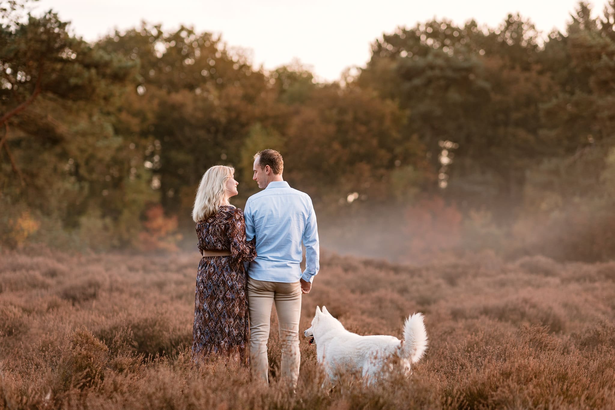 Man en vrouw staan samen met hun witte hond met hun rug naar de camera op de heide in de herfst.