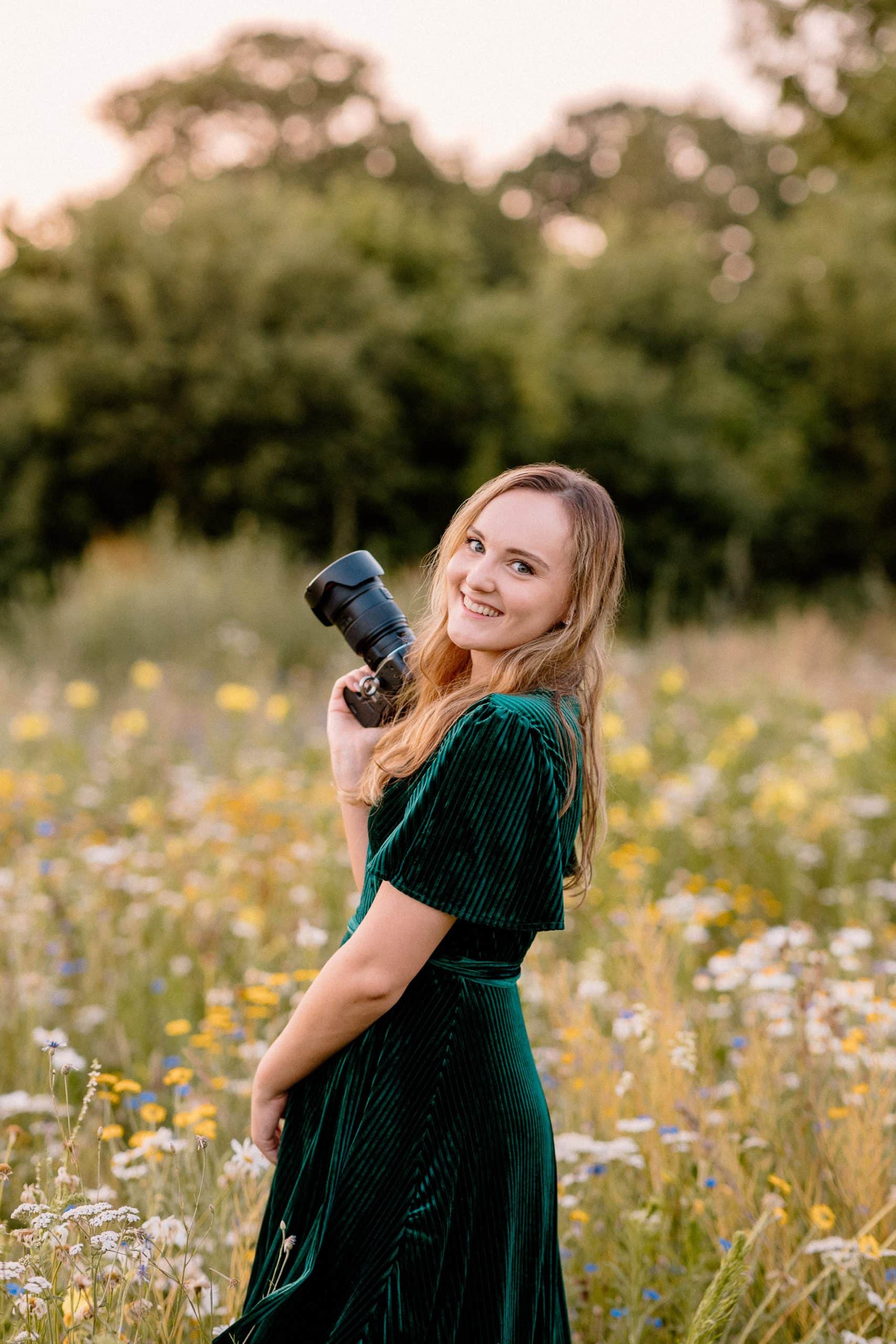 Fotograaf in een groene jurk staat met haar camera lachend in een bloemenveld.