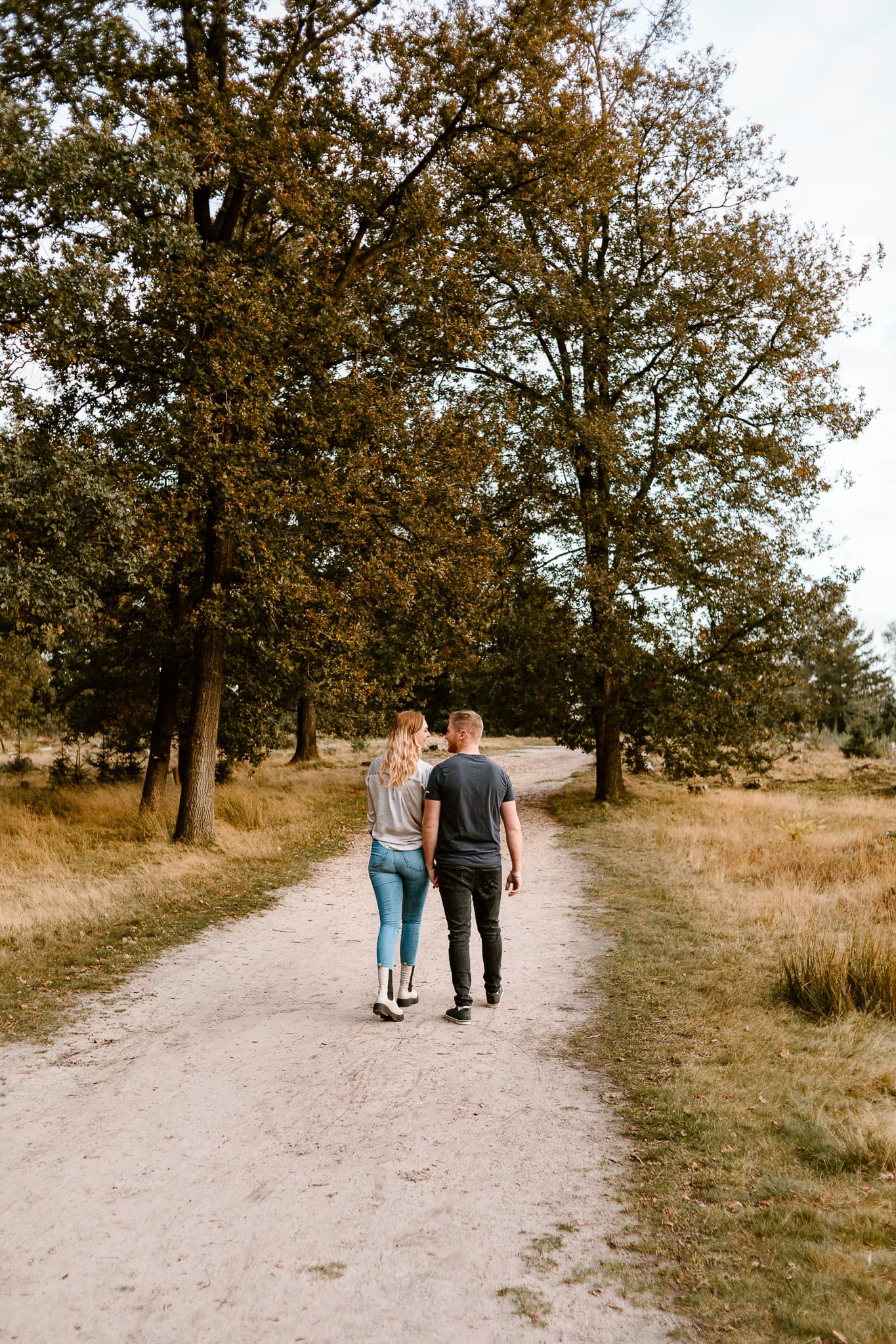 Man en vrouw in spijkerbroek lopen hand in hand over een bospad weg van de camera tijdens een loveshoot in Appelscha.