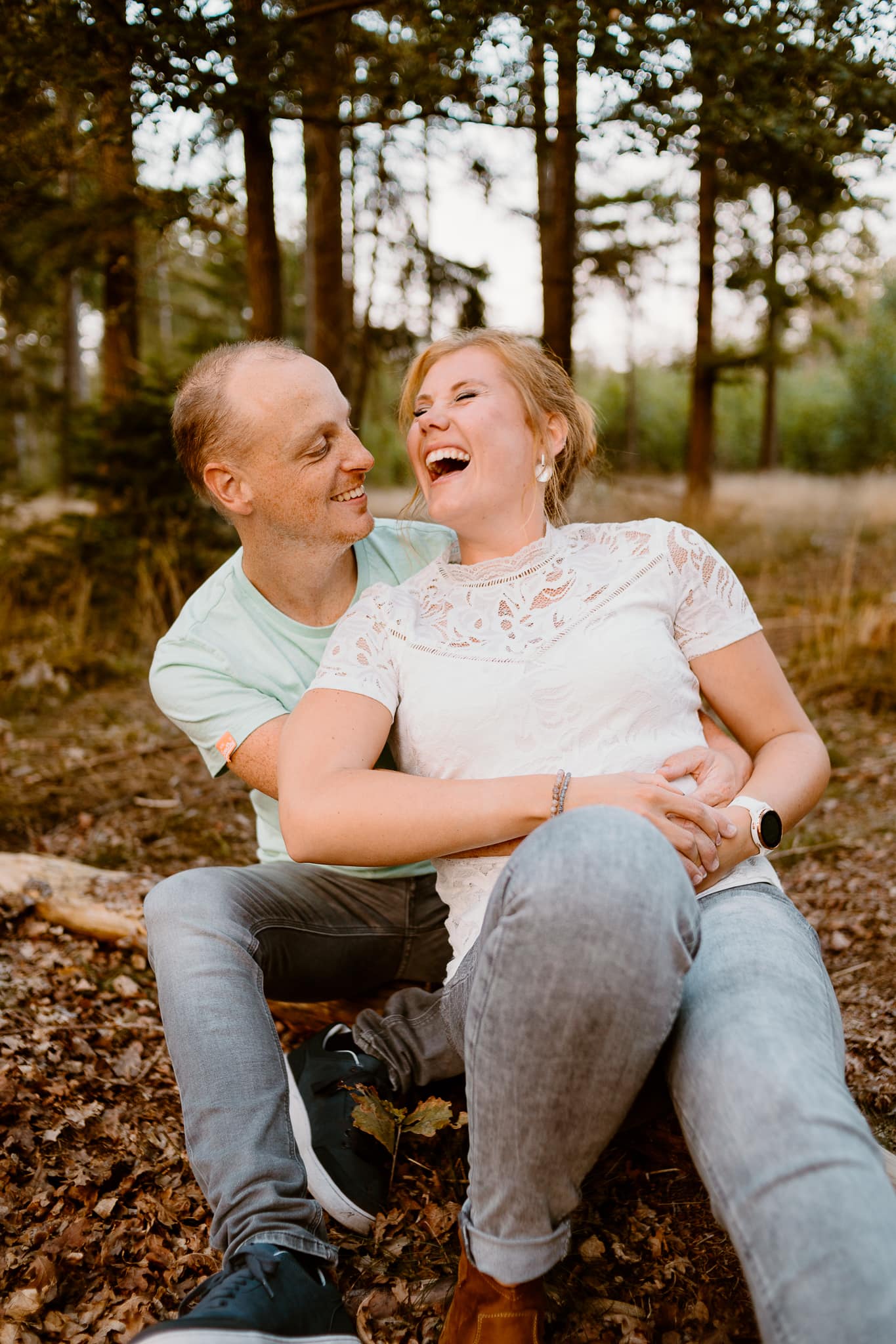 Vrouw in witte kanten top leunt lachend tegen haar man aan terwijl ze op de grond zitten tijdens een koppelshoot in het bos in Ees.