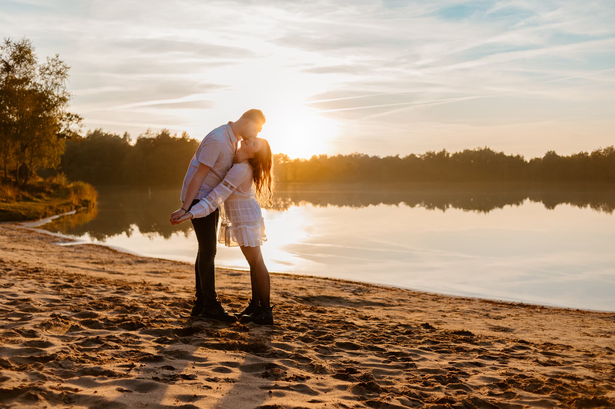 Meisje in witte jurk krijgt kus op haar wang van jongen in spijkerbroek en blouse op het strand van het Blauwe Meer in Dwingeloo met een prachtige zonsondergang op de achtegrond.