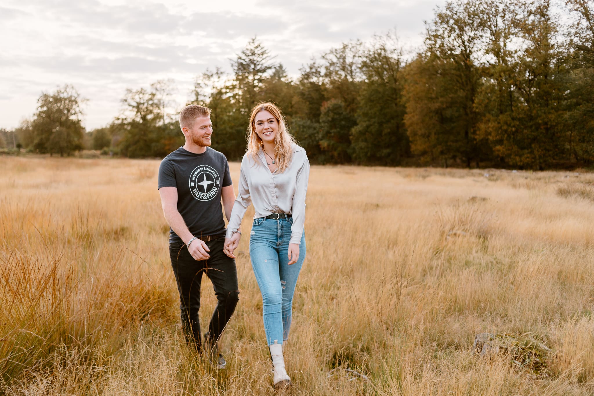 Koppel loopt hand in hand richting de camera tijdens een fotoshoot. Man kijkt lachend naar zijn vriendin.