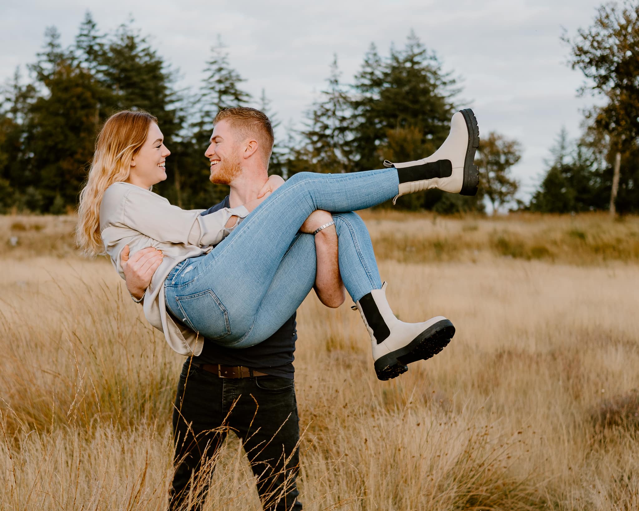 Jongen heeft zijn vriendin tijdens een fotoshoot in Friesland in beide armen opgetild. Zij zwaait haar benen in de lucht en ze kijken elkaar lachend aan.