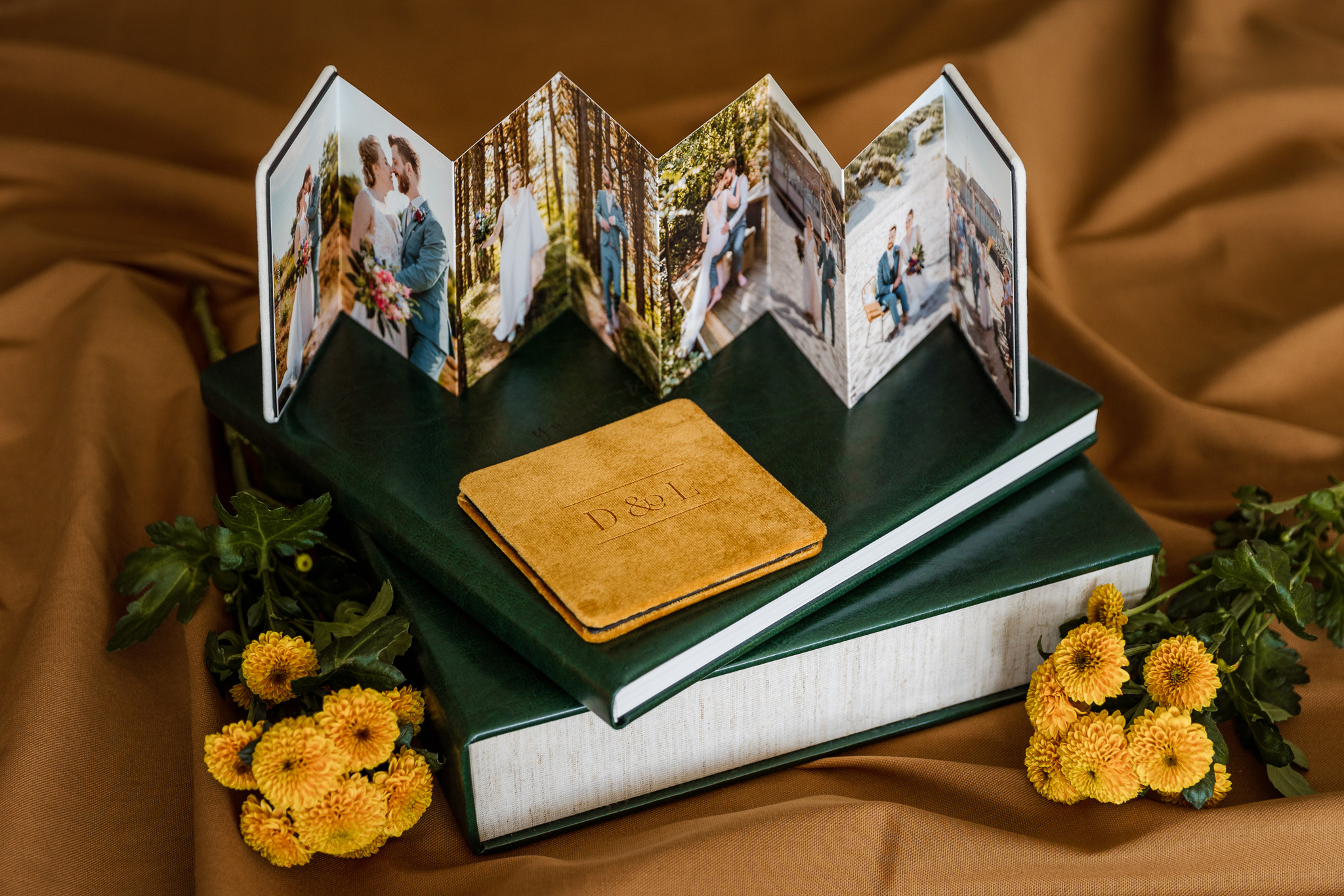 Een handgemaakt accordeonfotoboek gevuld met trouwfoto's, geplaatst op een stapel hardcoverboeken, omgeven door gele bloemen op een gedrapeerde bruine stof.