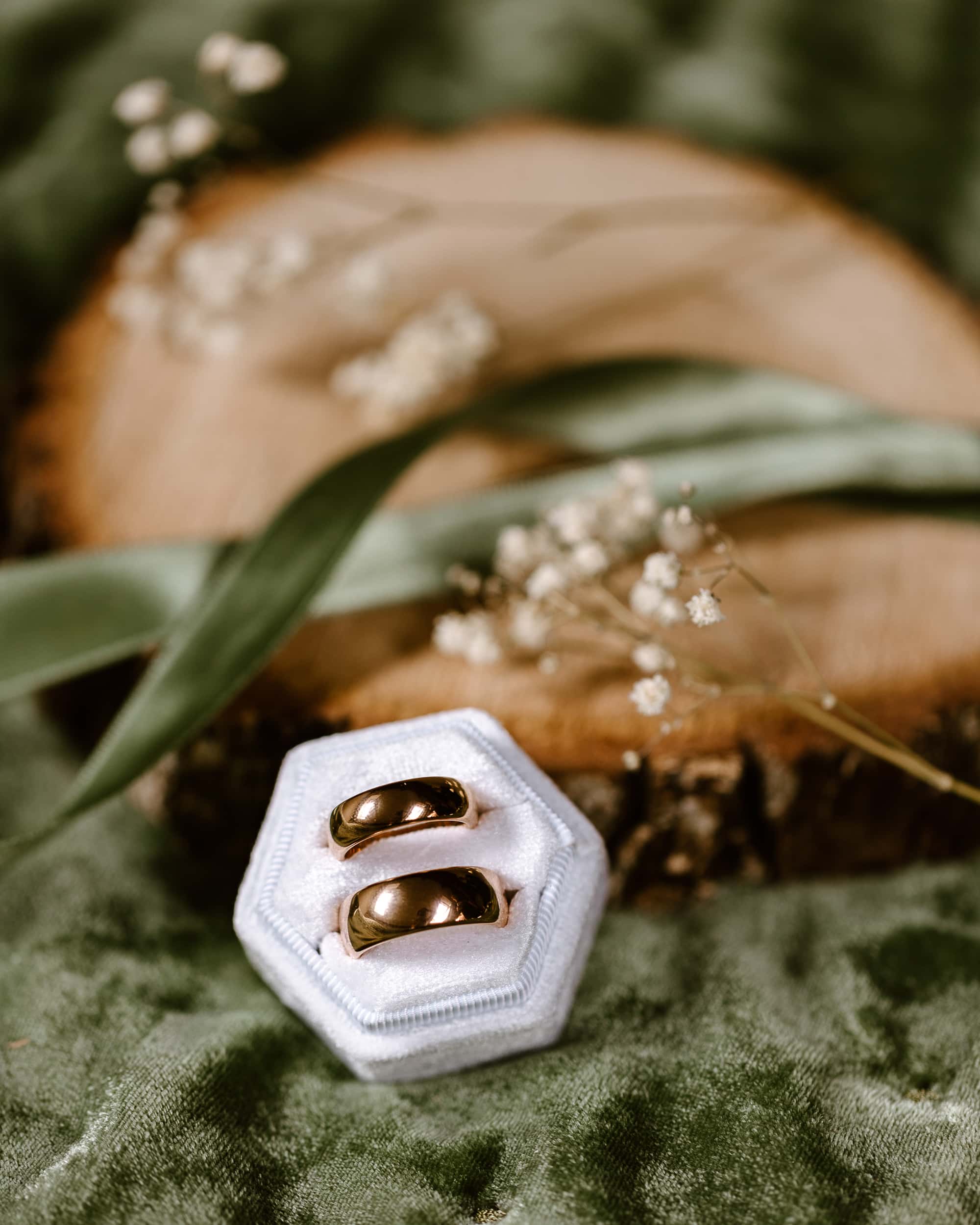 Twee trouwringen liggen in een wit ringendoosje op een groen bed met witte bloemetjes en groen lint op de achtergrond.